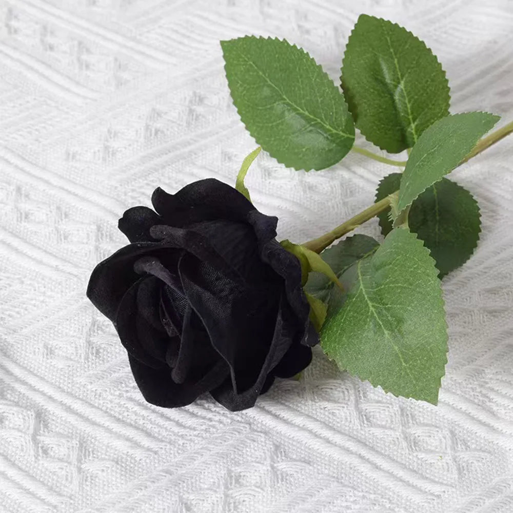 A single, dark black artificial rose with green leaves on a white textured background.