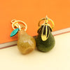 A gold-toned pear-shaped fruit keychain charm with a green gemstone hanging from it, set against an orange background.