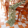 Sage green gauze table runner draped on a wooden dining table set for an event.