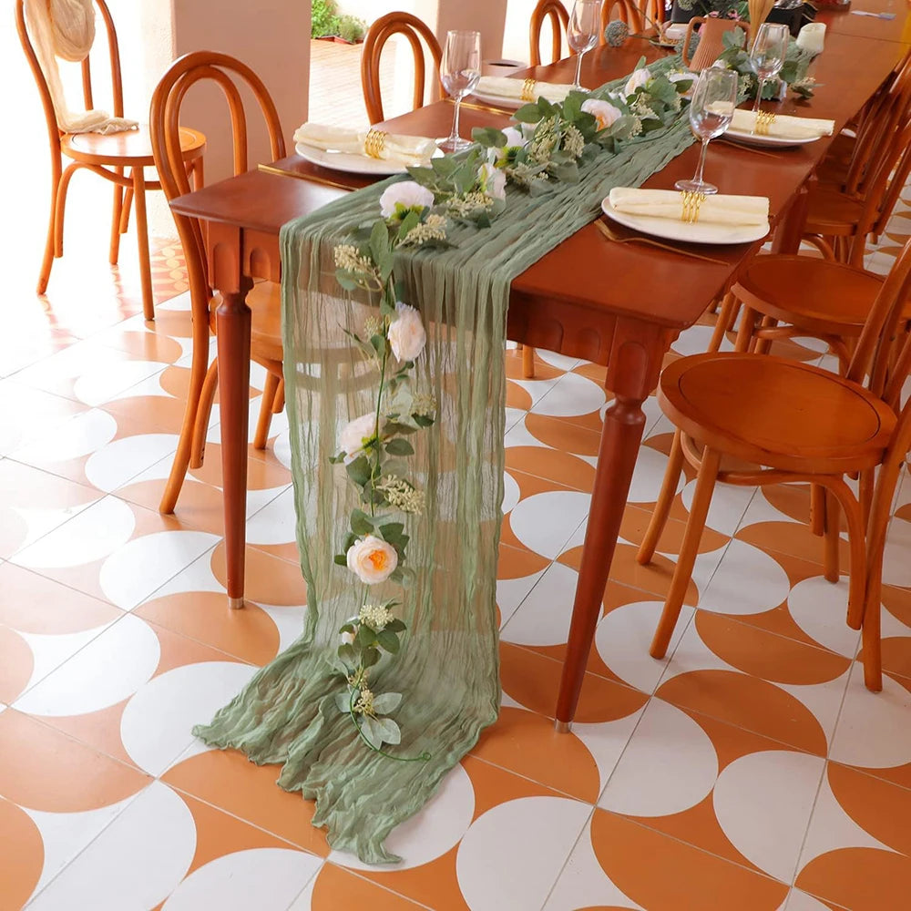 Sage green gauze table runner draped on a wooden dining table set for an event.