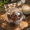 A clear plastic container filled with dark chocolate cake pieces sits on a wooden table next to a latte with latte art.