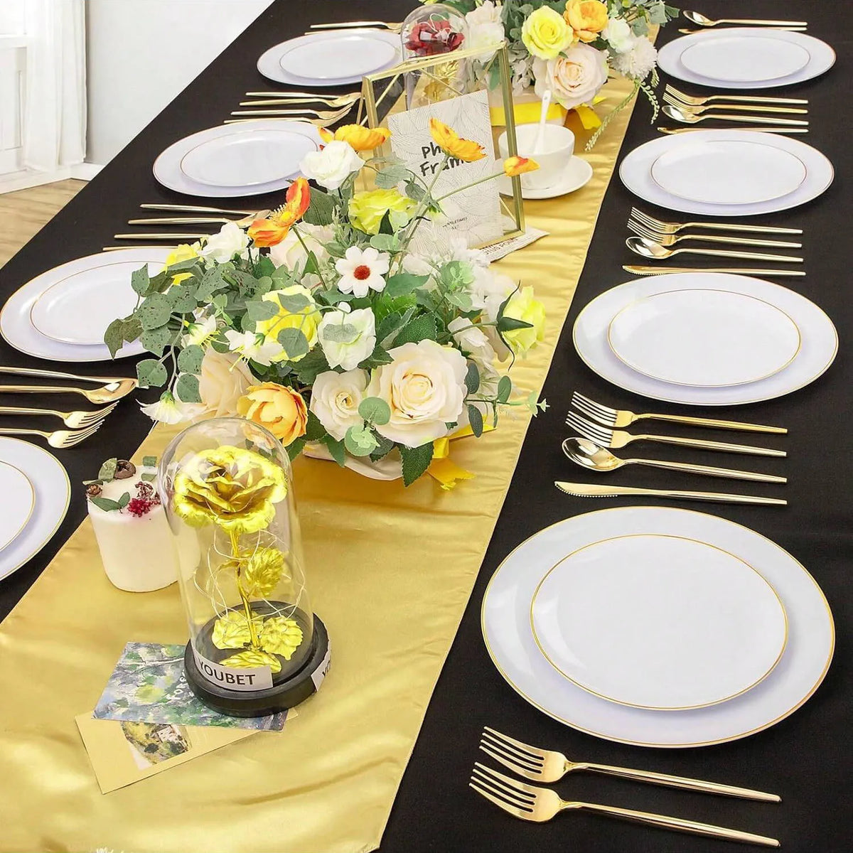 A display of gold rim plastic plates for weddings and parties set on a black tablecloth with floral arrangements.
