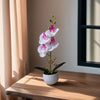 A realistic artificial orchid potted plant with white and purple flowers sits on a wooden table.