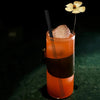 An orange-colored drink in a glass with a large ice sphere and a black straw, set against a dark background.