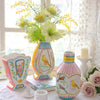 A ceramic bird floral vase with yellow and white patterns sits on a white lace cloth next to a window.