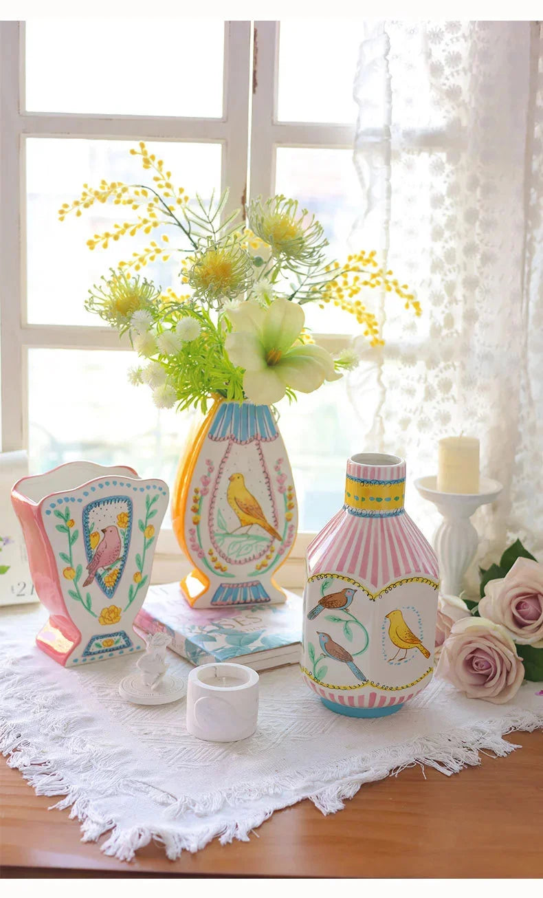 A ceramic bird floral vase with yellow and white patterns sits on a white lace cloth next to a window.
