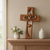A wooden wall cross with intertwined hearts rests on a light wood shelf next to a white ceramic vase.