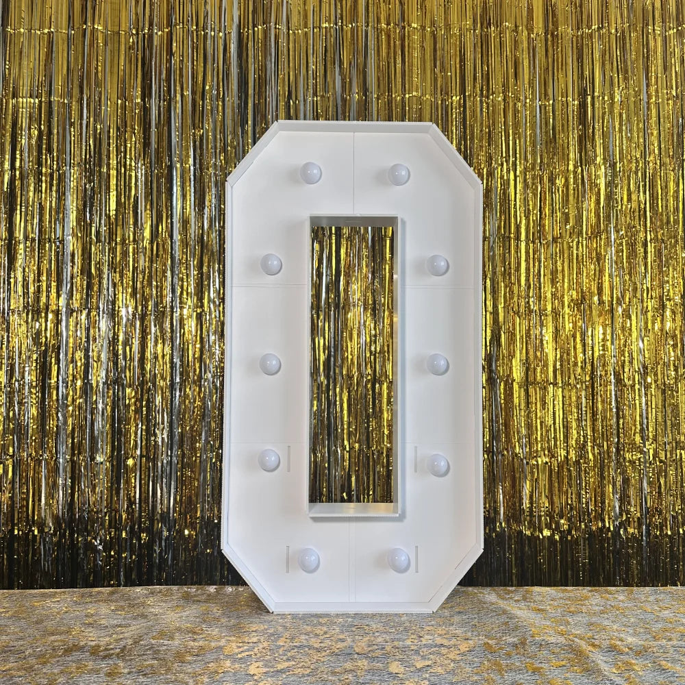 A large, white marquee light-up letter in the shape of the number '0' mounted against a shimmering gold curtain backdrop.