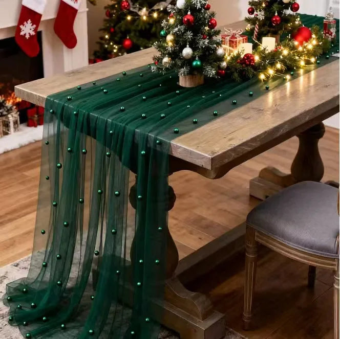 A wooden table draped with a sheer green fabric featuring beaded trim and festive holiday decorations.
