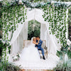 A bride and groom stand under a white archway decorated with greenery and flowers for a wedding photo.