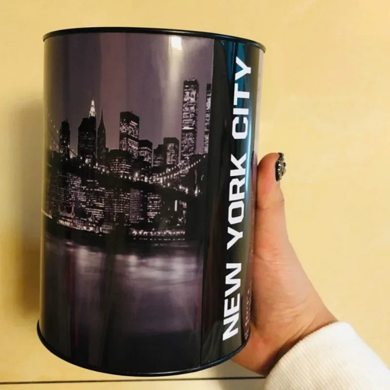 A hand holds a cylindrical metal piggy bank with a silhouette of a city skyline printed on it.
