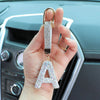 A crystal letter keychain charm hanging from a gold ring, held in a hand against a car interior.