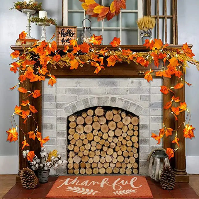 An autumn leaf garland with LED lights is displayed on a fireplace mantel decorated with fall foliage and firewood.