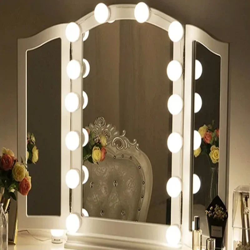 A white framed vanity mirror with round LED lights reflecting a silver chair and floral arrangements.