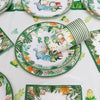 A green and white dinosaur-themed disposable tableware set featuring plates, a banner, and balloons on a white tablecloth.
