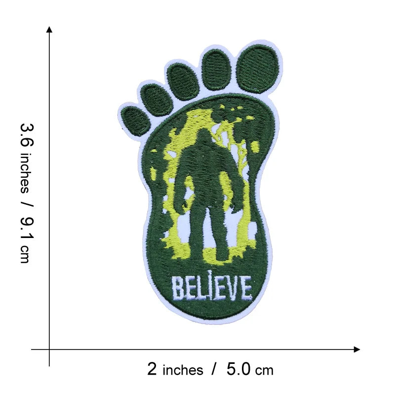 An embroidered patch shaped like a large green footprint with a yellow silhouette inside and the word BELIEVE below.