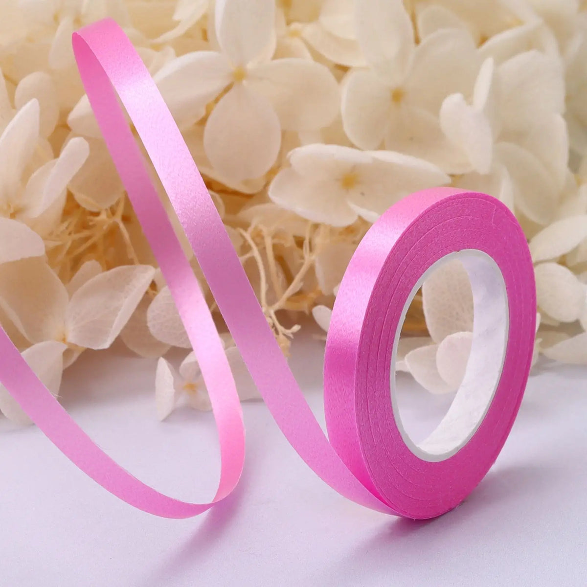 Pink satin balloon ribbon roll unwinding against a background of white flowers.