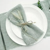 A stack of ten handcrafted fringed cotton napkins in a muted sage green color placed on white plates and a gray linen napkin.