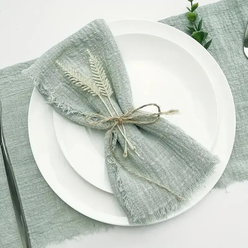 A stack of ten handcrafted fringed cotton napkins in a muted sage green color placed on white plates and a gray linen napkin.
