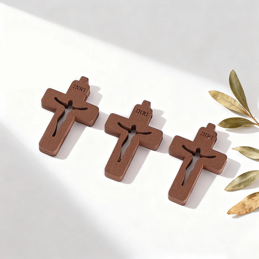Trois petits pendentifs en bois avec INRI gravé, posés sur une surface blanche à côté de feuilles d'olivier.