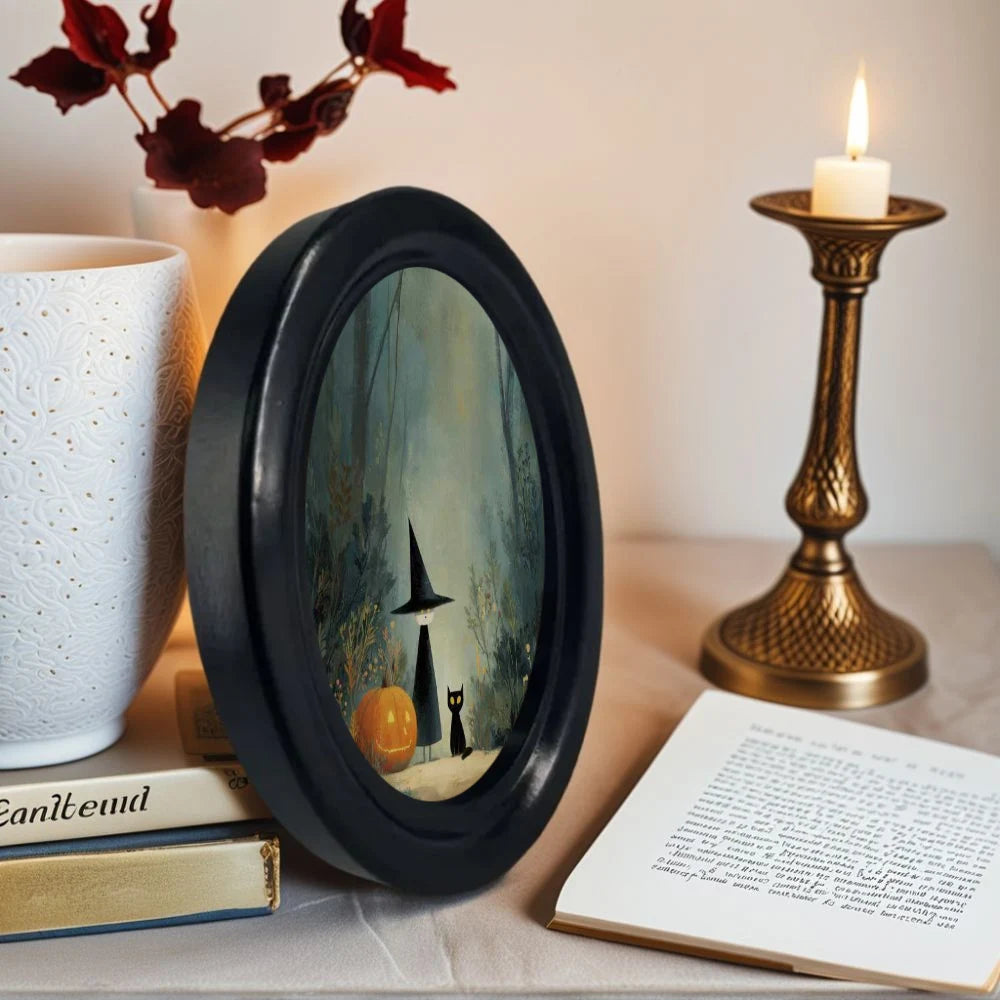 An oval framed piece of wall art featuring a witch and a black cat in a woodland scene
