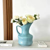 A light blue ceramic tabletop vase filled with white and yellow roses sits on a white shelf.