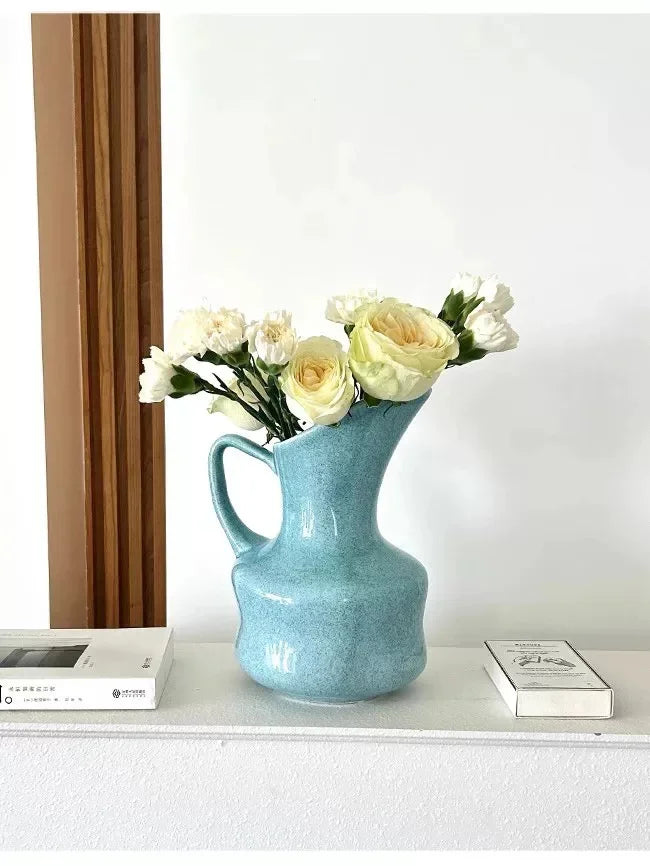 A light blue ceramic tabletop vase filled with white and yellow roses sits on a white shelf.