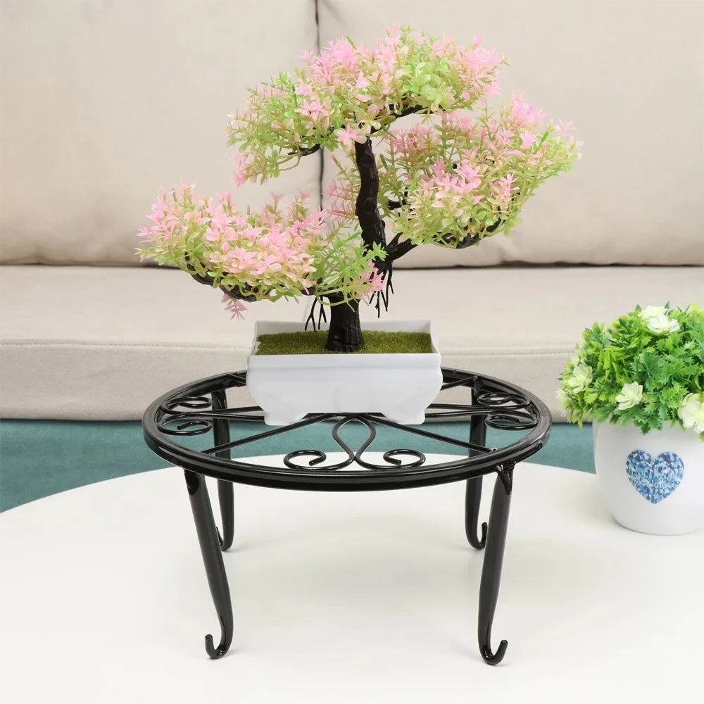 A decorative black metal plant stand with a saucer holding a small pink and green bonsai-style tree.
