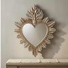 Ornate flame wall mirror with a heart shape framed in gold-toned metal hanging on a light gray wall above a wooden surface.