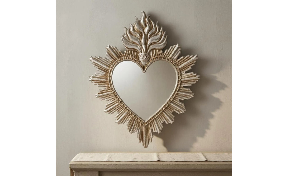 Ornate flame wall mirror with a heart shape framed in gold-toned metal hanging on a light gray wall above a wooden surface.
