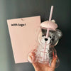 A hand holds a clear glass coffee cup shaped like a bear with a handle and a pink straw against a light pink card.