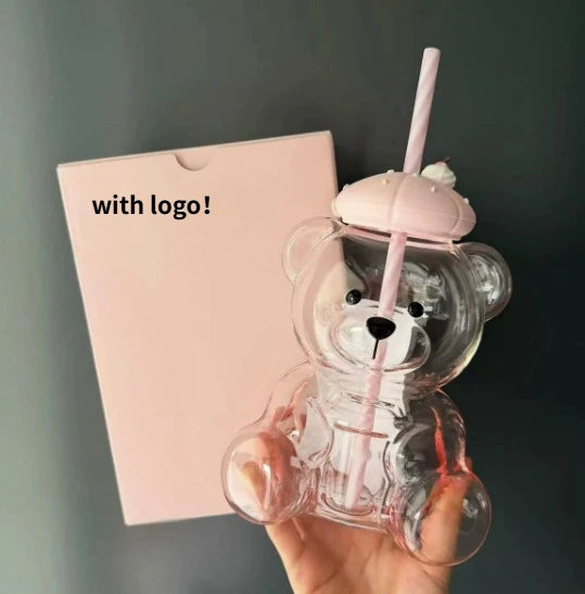 A hand holds a clear glass coffee cup shaped like a bear with a handle and a pink straw against a light pink card.