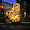 A solar-powered owl garden stake light illuminates a realistic owl sculpture in the evening setting.