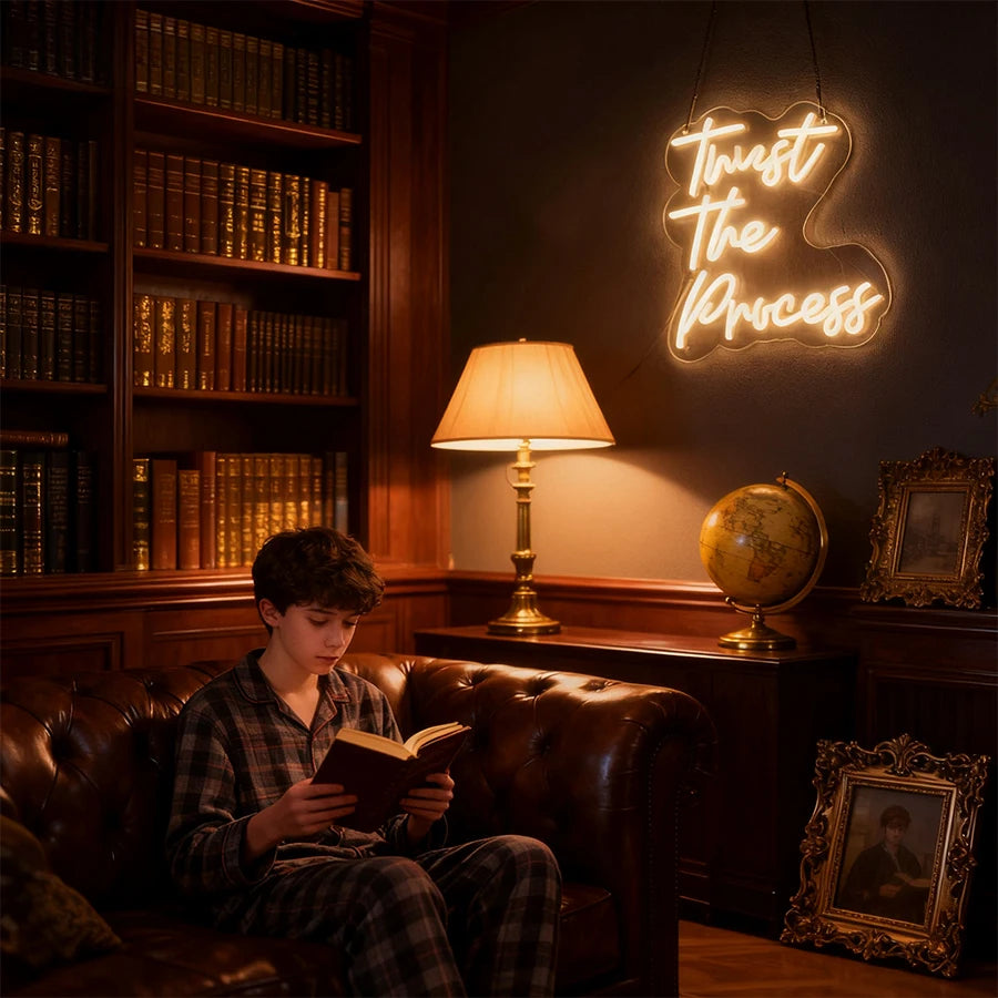 A young man sits in a leather armchair reading a book in a dimly lit room with a glowing neon sign on the wall.