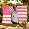 A man stands in front of a pink and white American flag tapestry hanging outdoors.
