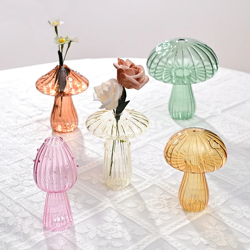 Collection de vases décoratifs en champignons de verre dans des tons rose, vert et ambre sur une nappe en dentelle blanche.