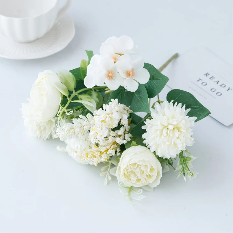 A bouquet of white artificial silk peony flowers with green leaves and a small card on a white surface.