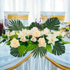 A floral arrangement featuring peach and cream roses and large green tropical palm leaves on a white tablecloth.