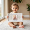 A baby is holding a white frame containing her handprint and footprint, sitting on a light rug.