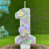 A tall candle shaped like the number one decorated with purple glitter, a white flower, and a light purple butterfly on a woo