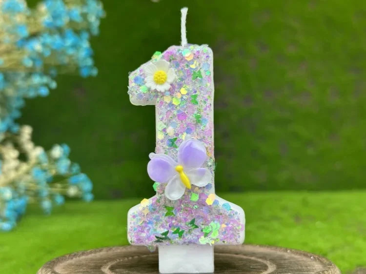 A tall candle shaped like the number one decorated with purple glitter, a white flower, and a light purple butterfly on a woo