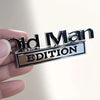 A hand holds a silver metal emblem with the text Old Man Edition engraved on it.