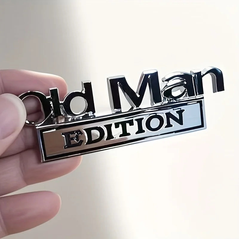 A hand holds a silver metal emblem with the text Old Man Edition engraved on it.