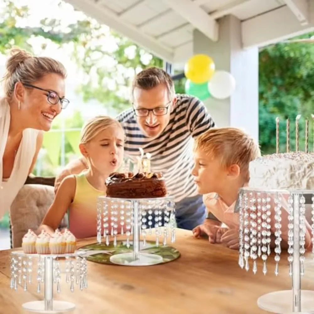 A family is gathered around a wooden table displaying an elegant transparent acrylic cake display rack with small cupcakes.