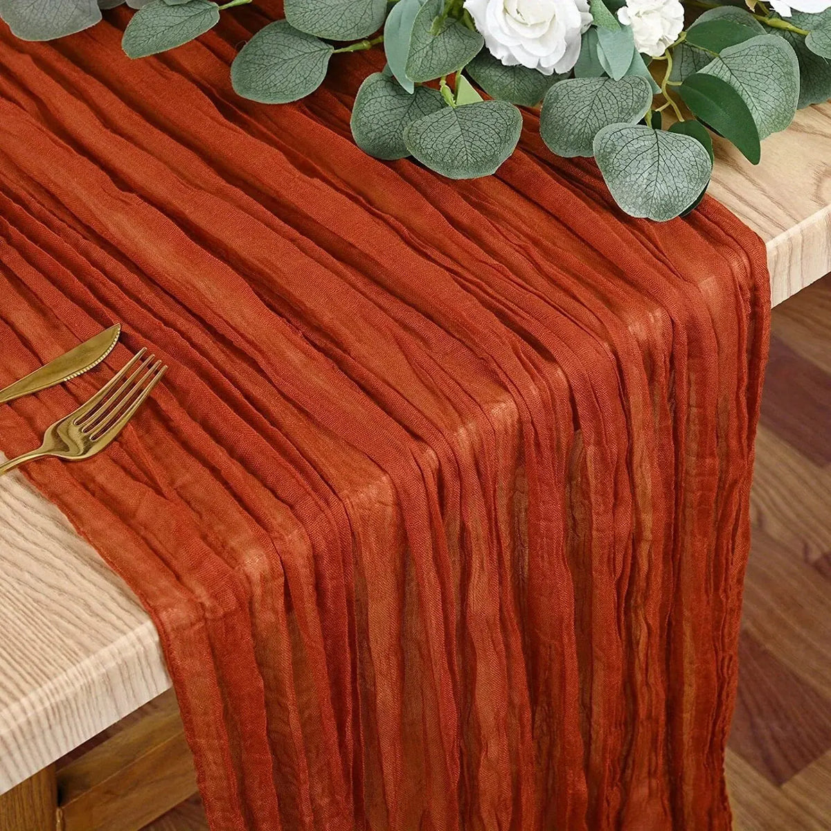 A rust orange boho style cheesecloth table runner draped over a wooden table with white flowers and a gold fork.