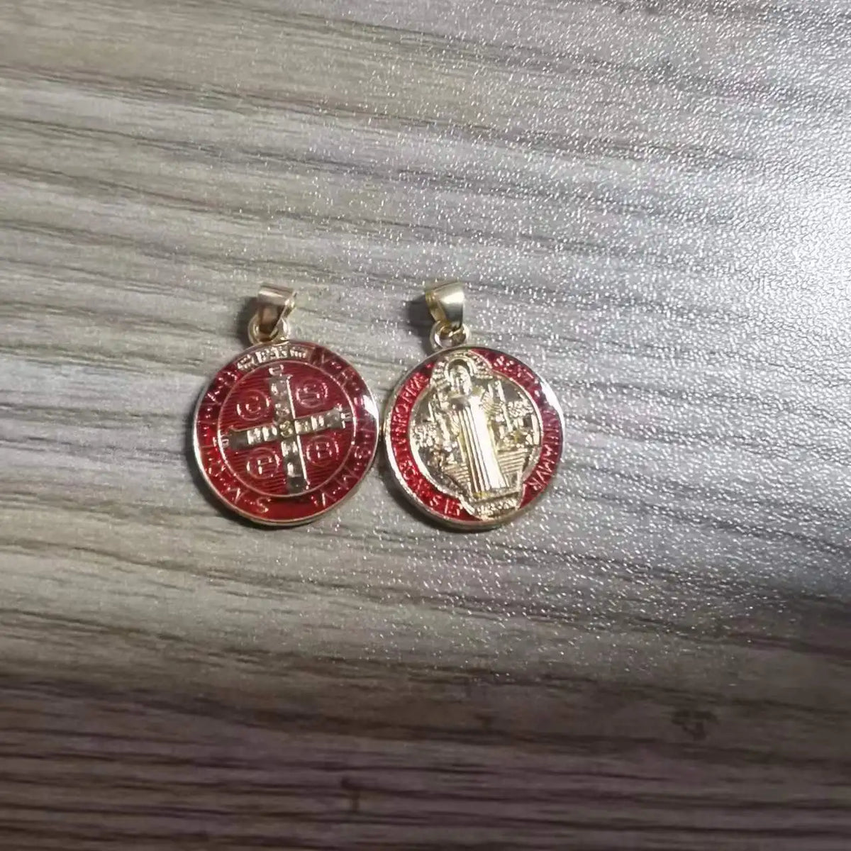 A pair of gold-toned religious pendants featuring a cross and a depiction of St. Benedict on red circular medallions.