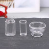 A set of clear glass containers including a bottle, a tall cylinder, and a small bowl, displayed on a dark surface.