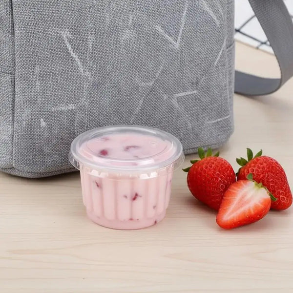 A clear disposable dessert cup filled with pink strawberry smoothie sits on a wooden surface next to fresh strawberries.