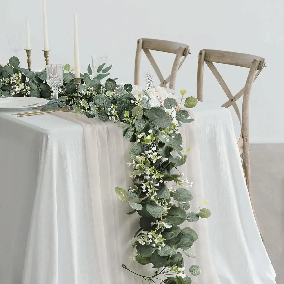 A long garland of green eucalyptus and white flowers decorates a white draped table with wooden chairs.