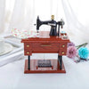 A miniature black sewing machine with a gold detail sits on a wooden box, set against a white background.